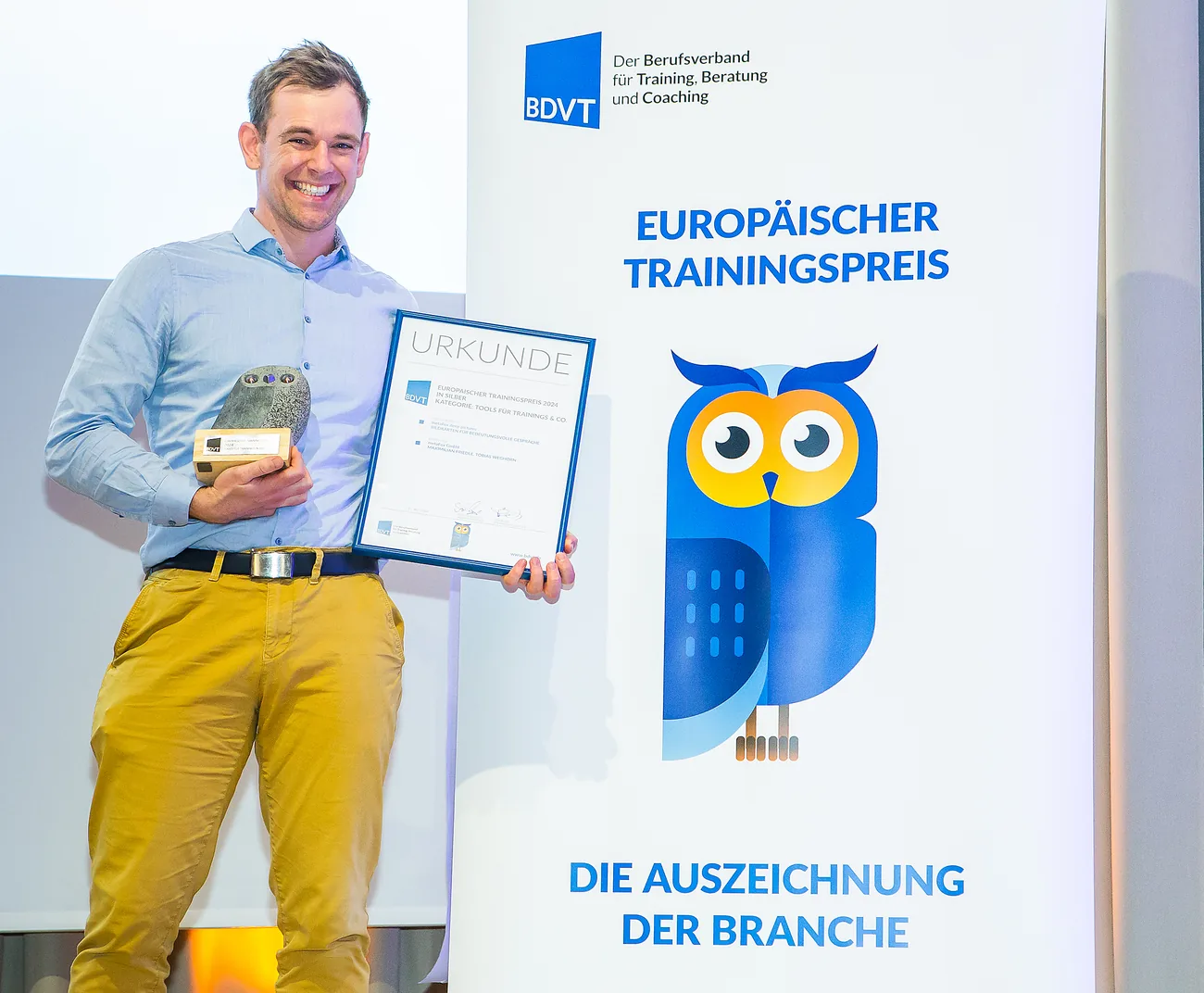 Max Friedle on stage holding the European Training Award certificate and trophy in front of the BDVT banner
